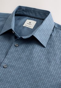 Chemise bleu clair coupe régulière avec un motif géométrique texturé, col boutonné et un bouton près de la poitrine. Étiquette incluse à l'intérieur.