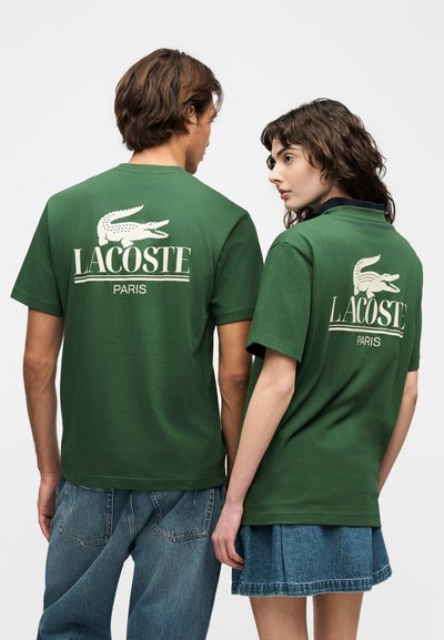 Двама души, носещи зелени тениски Lacoste Paris с бели крокодилски лога на гърба, стоящи един до друг с гръб към зрителя.