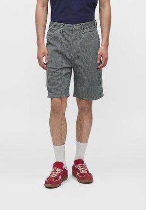 Homme portant un short rayé vertical noir et blanc, des chaussettes blanches montantes, des baskets rouges et une chemise bleu marine visible à la taille.