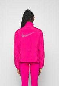 Różowa kurtka Nike z wysokim kołnierzem, mankietami i odblaskowym logo na plecach. Gładki, błyszczący materiał z luźnym fasonem.