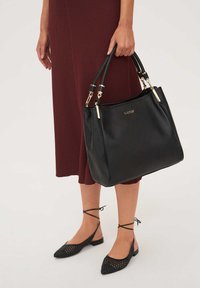 Melnās ādas rokassomiņa ar akmeņainu tekstūru, zelta aparatūras akcentiem, divām rokturām un plašu dizainu, ko tur cilvēks.