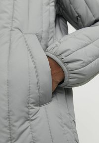 Giubbotto imbottito grigio con design trapuntato, dotato di tasca sul lato sinistro e polsini elasticizzati per un look aderente. Texture liscia.