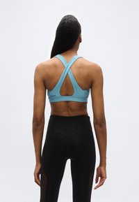 Γυναίκα από πίσω φορώντας ένα μπλε αθλητικό σουτιέν με σταυρωτές τιράντες και μαύρο κολάν με ψηλή μέση, μπροστά σε απλό ανοιχτό φωτεινό φόντο.