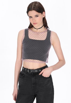 Giovane donna che indossa un top corto nero trasparente a pois sopra una canottiera nera, jeans neri a vita alta e una collana nera con ciondolo.