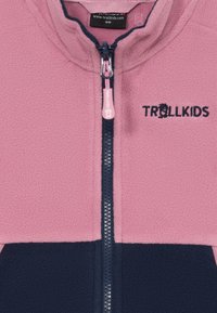 Roza polarna jakna z zadrgo spredaj, mornarsko modri detajli na dnu in roza zadrgo. Ima vezen logo "TROLLKIDS" v mornarsko modri barvi.