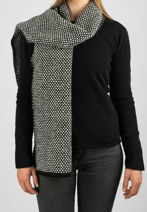 Kobieta ubrana w czarną bluzkę z długim rękawem i ciemne dżinsy, z luźno owiniętym wokół szyi czarno-białym teksturowanym szalikiem.