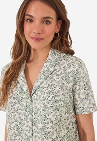Chemise de pyjama à manches courtes en tissu léger, ornée de feuilles vertes et de fleurs jaunes sur fond blanc, avec une poche poitrine et une fermeture à boutons.