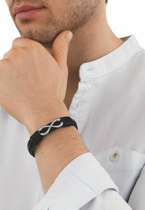 Primer plano de una persona con una camisa blanca, apoyando la barbilla en la mano, mostrando una pulsera de cuero negro con un símbolo de infinito plateado.