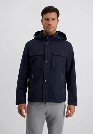 Man draagt een marineblauw joggingjack met capuchon, voorzien van klepzakken aan de voorkant en ritssluitingen, gecombineerd met lichtgrijze broek, staand tegen een effen achtergrond.