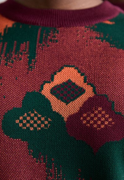 Sweter robiony na drutach z okrągłym dekoltem. Charakteryzuje się zieloną, bordową i pomarańczową paletą kolorów oraz geometrycznymi i kwiatowymi wzorami. Teksturowana powierzchnia.