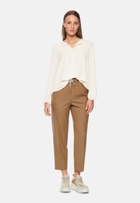Hellbeige Bluse mit Kordelzugkapuze, kombiniert mit braunen, taillierten Hosen und beigen Sneakers, die über strukturierte Details und einen lässigen Schnitt verfügen.