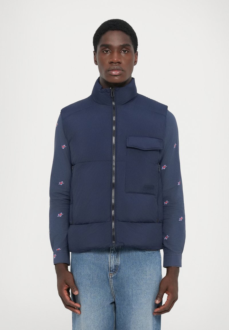 Navy blue puffer vest with high collar, front zip, and a chest pocket. Features patterned long sleeves with small floral designs.
