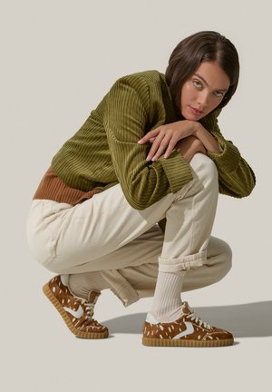 Brown sneaker with white accents, textured upper, round toes, and rubber sole. Worn with light-colored pants and ribbed socks.