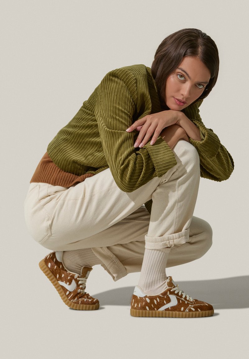 Brown sneaker with white accents, textured upper, round toes, and rubber sole. Worn with light-colored pants and ribbed socks.