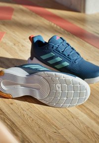 Un paio di scarpe da ginnastica blu con strisce azzurre e suole bianche su un pavimento in legno, una scarpa mostra la suola testurizzata.