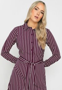 Abito camicia prugna con strisce verticali bianche, maniche lunghe, apertura frontale con bottoni e una cintura annodata in vita. Presenta un colletto e una tasca sul petto.