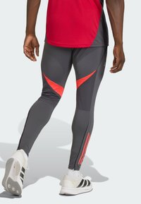 Γκρι αθλητικό κολάν με κόκκινες γωνιώδεις λεπτομέρειες, με εφαρμοστή σχεδίαση και φερμουάρ στα τελειώματα. Φορεμένο με λευκά αθλητικά παπούτσια με μαύρες ρίγες.