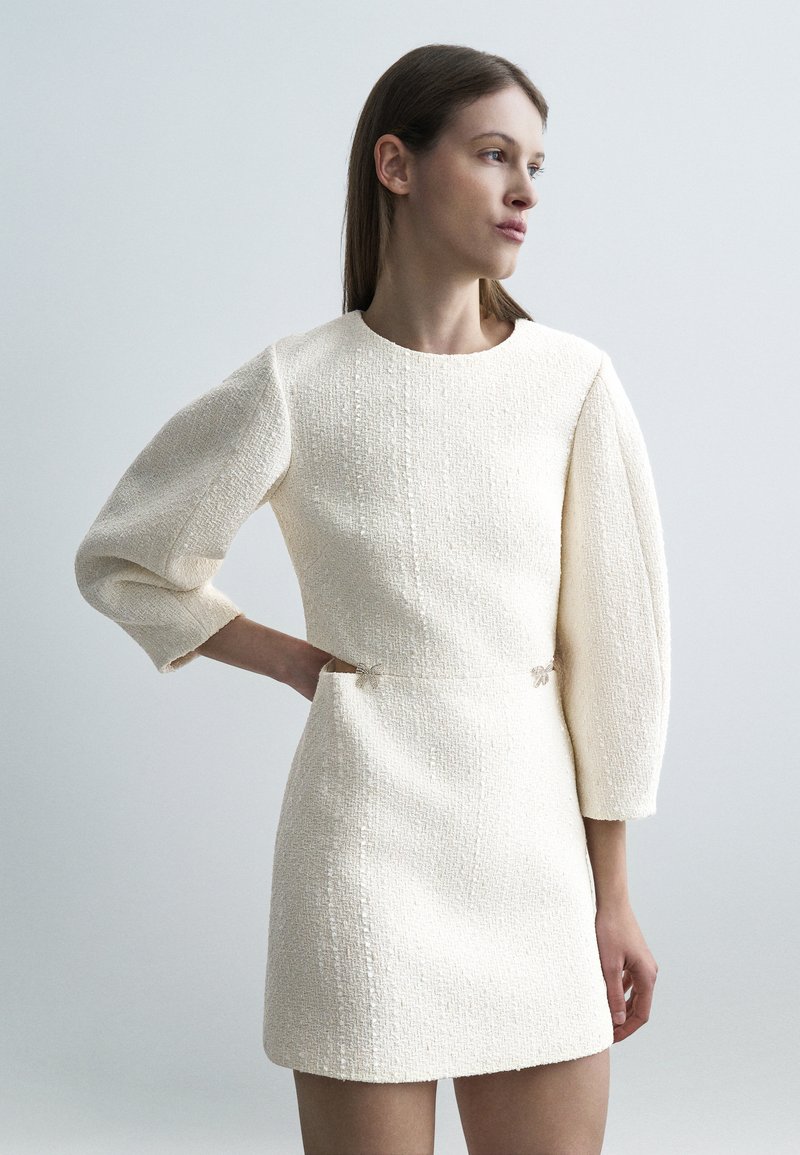 Γυναίκα που φοράει ένα υφασμάτινο, εκρού φόρεμα με φουσκωτά μανίκια τριών τετάρτων, στέκεται με το ένα χέρι στη μέση, κοιτάζοντας προς τα πλάγια μπροστά σε ένα απλό φόντο.