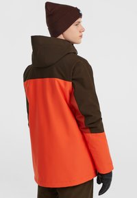 Multikleurige waterdichte jas in oranje en bruin, met een capuchon en aansluitende manchetten. De achterkant heeft een kleurblokontwerp. Zwarte handschoenen zichtbaar.