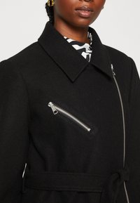 Abrigo de lana negro con un cuello afilado, cintura ajustada con cinturón y un bolsillo en el pecho con cremallera en ángulo y herrajes plateados. Tejido suave y texturizado.
