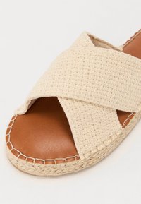 Beige vevd slip-on sandal med kryssede tekstilstropper, brun skinnsåle og vevd jutefot. Søm detaljer langs kanten.