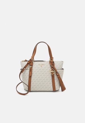 White canvas tote bag with gray "MK" logo pattern, brown leather accents, double handles, detachable shoulder strap, and tassel detail.