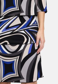 Kleid mit geometrischem Muster in Schwarz, Grau und Blau. Weicher Stoff mit geschichtetem Design und schmaler Silhouette. Hand auf dem Rock platziert.