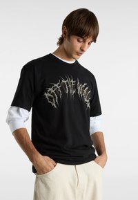 Sort t-shirt med et stort sølv grafisk design, rund halsudskæring og korte ærmer, lagdelt over en hvid langærmet skjorte.
