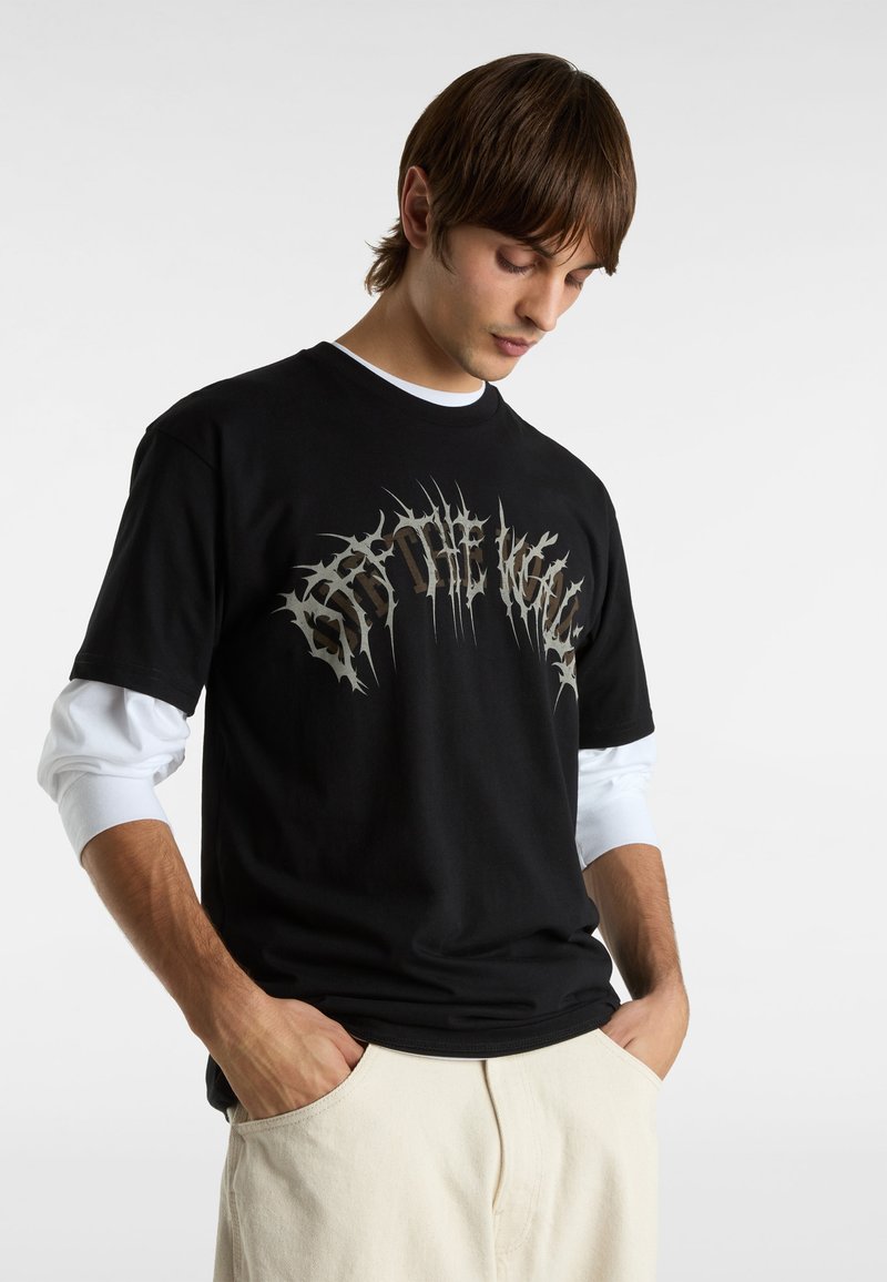 Zwart t-shirt met een groot zilver grafisch ontwerp, ronde halslijn en korte mouwen, gedragen over een wit langemouwen shirt.