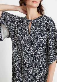 Marineblaues Kleid mit cremefarbenem Blumendruck, kurzen schwingenden Ärmeln, Schlüssel-Ausschnitt und weichem Stoff, der eine lockere, fließende Passform bietet.