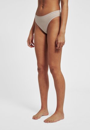 Kobieta stoi boso, nosząc beżowe majtki z wysokim stanem na jednolitym białym tle.