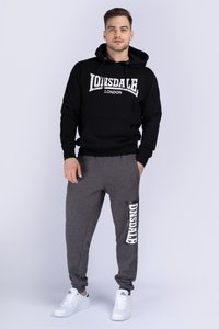 Sudadera con capucha negra con logo blanco "LONSDALE LONDON", combinada con pantalones de chándal grises que presentan una impresión blanca "ONSDALE" y puños elásticos. Zapatillas blancas.