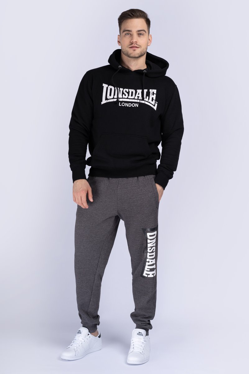Sudadera con capucha negra con logo blanco "LONSDALE LONDON", combinada con pantalones de chándal grises que presentan una impresión blanca "ONSDALE" y puños elásticos. Zapatillas blancas.