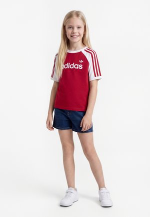 Usmievavé dievča s dlhými blond vlasmi, oblečené v červenom tričku Adidas, modrých rifľových šortkách a bielych teniskách, stojace na bielom pozadí.