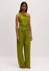 Groene een-schoudertop in combinatie met bijpassende wijde broek. Zachte stof, ontspannende pasvorm en geplooide details bij de taille. Platte sandalen maken de look compleet.