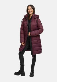 Mujer sonriendo, vestida con un abrigo acolchado largo burdeos con capucha, jeans negros y botas gruesas negras, de pie contra un fondo liso.