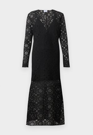 Femme portant une longue robe noire en dentelle avec des motifs floraux, portant un grand sac à épaule marron et des chaussures plates noires.