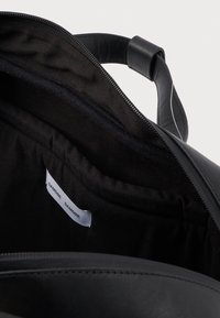 Sac en cuir noir avec fermeture éclair, intérieur texturé et une étiquette à l'intérieur. Dispose d'une poignée arrondie pour le transport.