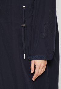 Coupe-vent bleu marine avec un poignet texturé, un cordon élastique avec des basculeurs, et un détail en texte en relief sur la manche. Finition en tissu lisse.