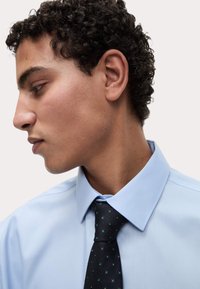 Jasnoniebieska koszula z wyraźnym kołnierzykiem, zestawiona z ciemnym krawatem w zielone kropki, prezentująca gładką fakturę i dopasowany krój.