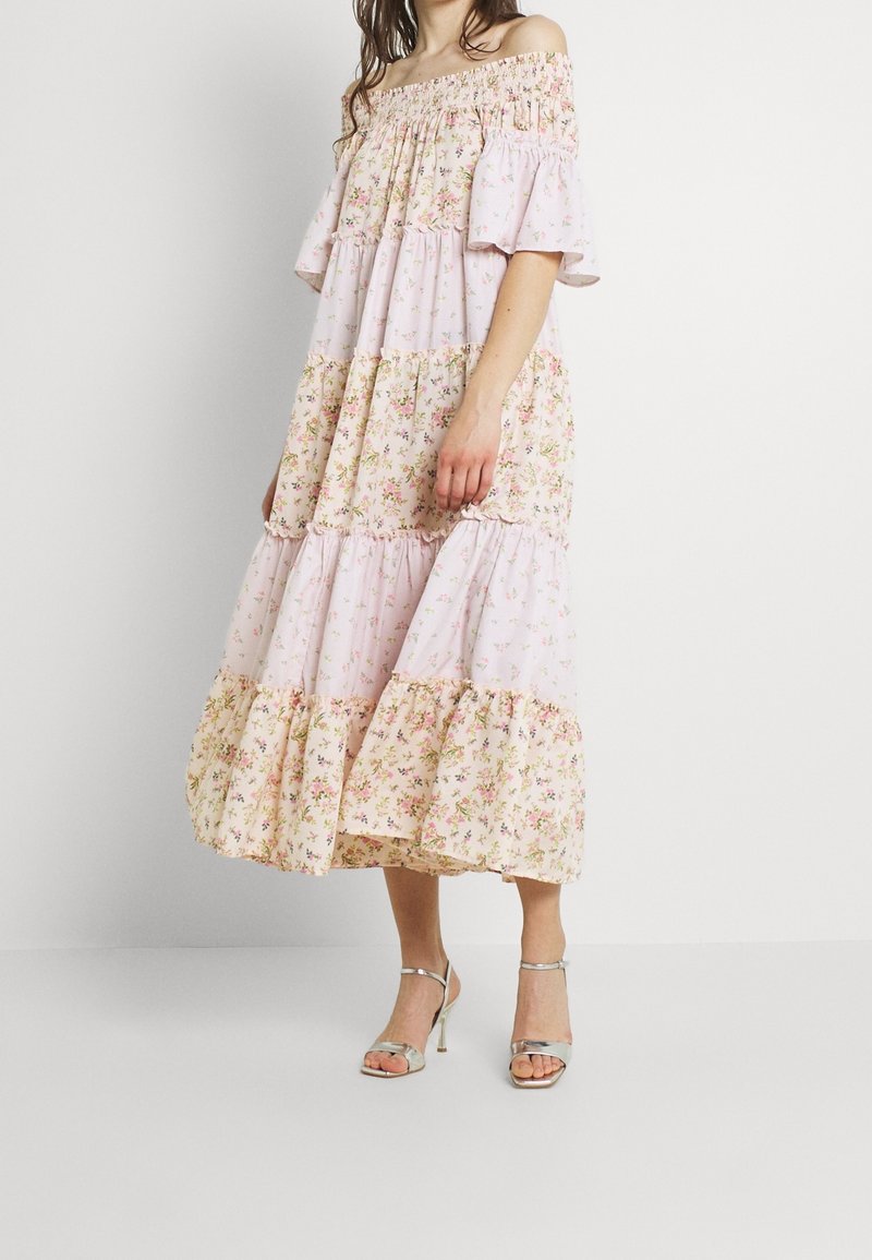 Femme portant une robe midi à volants fleurie rose et pêche, épaules dénudées, manches à volants et sandales à talons hauts argentées avec brides.