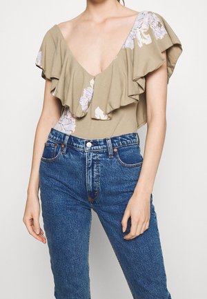Femme portant un jean bleu taille haute et un haut beige à fleurs avec un large détail volanté et un décolleté en V profond.