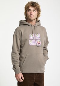 Sudadera marrón de tela suave con un bolsillo frontal. Presenta un gráfico de caras de dibujos animados en marcos rosas y el texto "VOLCOM".