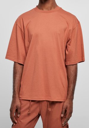 Homme portant un t-shirt ample couleur terre de Sienne et un pantalon à cordon assorti, debout devant un fond gris uni.
