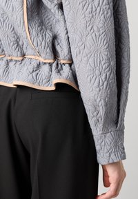 Gequilted lichtgrijze jas met bloemenpatroon, contrasterende perzikkleurige afwerking en lange mouwen, gecombineerd met zwarte broeken met een achterzak.