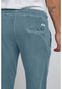 Lyseblå joggebukser lavet af blødt stof, med elastisk talje, sidelomme og diskrete syninger.