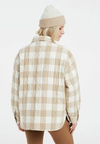 Vaatteena on pehmeä beige ja valkoinen ruutukuosiset vuoritettu takki, jossa on rento istuvuus, kärjellinen kaulus ja napitus. Asusteena käytetään neulottua beigenväristä pipoa.
