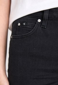 Zwarte spijkerbroek van denim met een klassiek ontwerp, voorzien van zilverkleurige hardware, een voorzak en een zwart etiket met witte tekst.