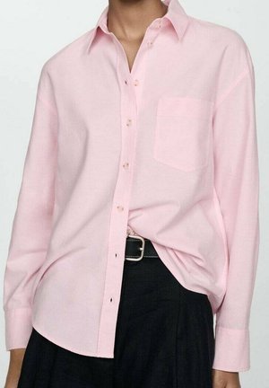 Osoba nosząca jasnoróżową koszulę na guziki z kieszenią na piersi, włożoną do czarnych spodni z czarnym paskiem z srebrną klamrą.