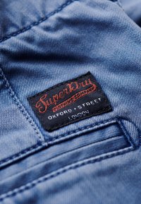 Tessuto di denim blu con una superficie tessurizzata. Presenta un'etichetta nera con "Superdry" in scritta rossa, che riporta "Oxford Street London."